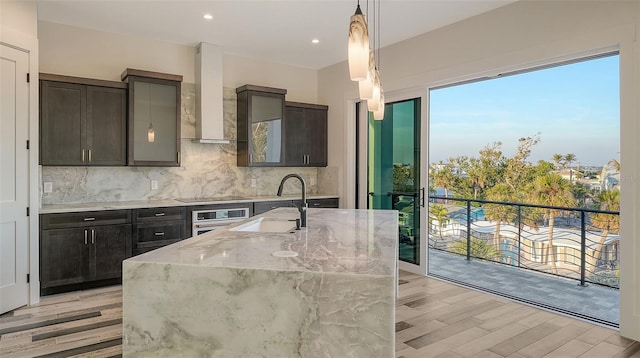 kitchen with sink, hanging light fixtures, stainless steel oven, light stone counters, and wall chimney exhaust hood