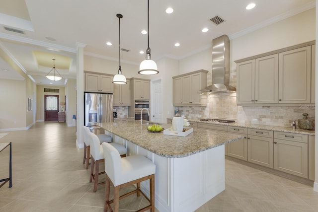 kitchen featuring light stone counters, decorative light fixtures, stainless steel appliances, a kitchen island with sink, and wall chimney range hood