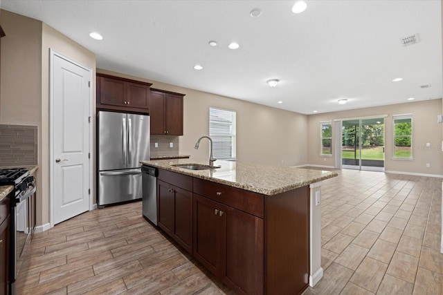 kitchen featuring sink, appliances with stainless steel finishes, light stone counters, an island with sink, and decorative backsplash