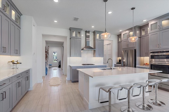 kitchen with a breakfast bar, appliances with stainless steel finishes, a kitchen island with sink, decorative light fixtures, and wall chimney exhaust hood