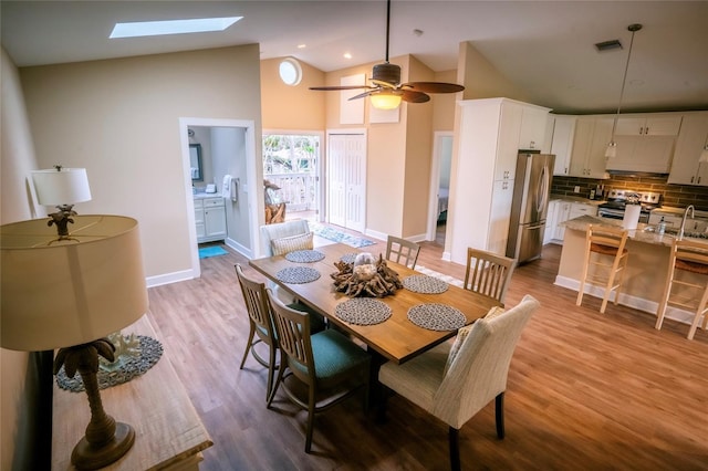 dining space featuring high vaulted ceiling, a skylight, sink, ceiling fan, and light wood-type flooring