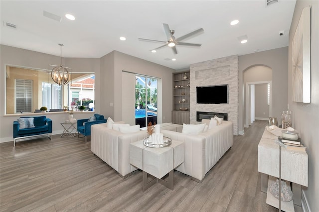 living room featuring built in shelves, a stone fireplace, ceiling fan with notable chandelier, and light hardwood / wood-style floors