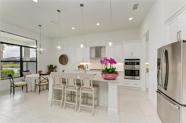 kitchen with an island with sink, white cabinets, hanging light fixtures, stainless steel appliances, and wall chimney exhaust hood