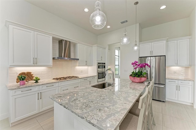 kitchen featuring a kitchen island with sink, a sink, white cabinetry, appliances with stainless steel finishes, and wall chimney exhaust hood