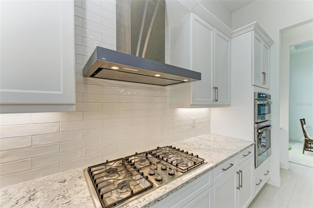 kitchen with tasteful backsplash, appliances with stainless steel finishes, white cabinets, wall chimney range hood, and light stone countertops