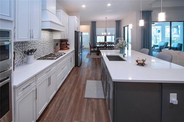 kitchen with appliances with stainless steel finishes, an island with sink, white cabinets, hanging light fixtures, and custom range hood