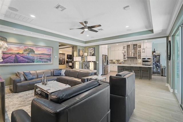 living room featuring a tray ceiling, visible vents, ornamental molding, light wood-style floors, and a ceiling fan