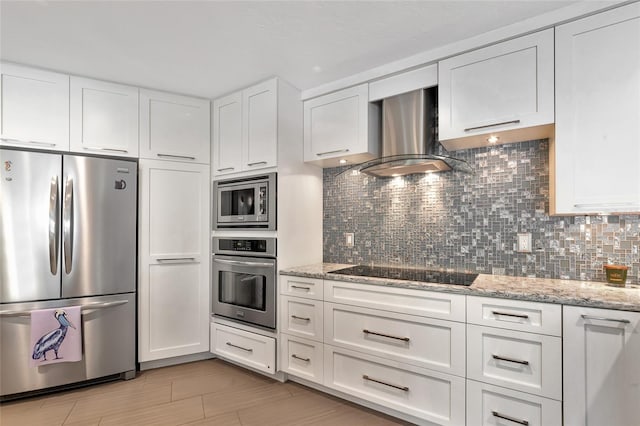 kitchen featuring stainless steel appliances, light stone counters, white cabinets, wall chimney range hood, and decorative backsplash