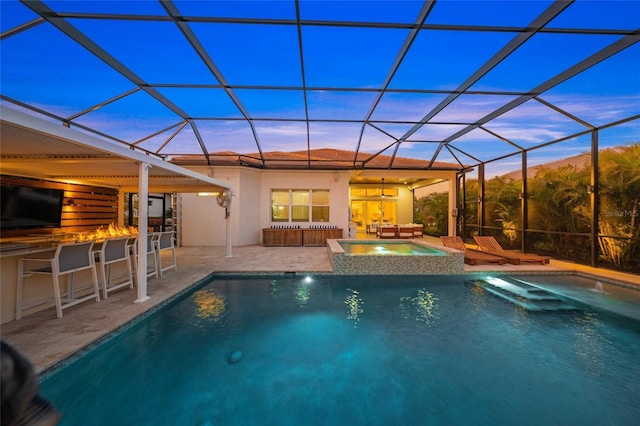 pool at dusk featuring an in ground hot tub, a patio area, an outdoor bar, and glass enclosure