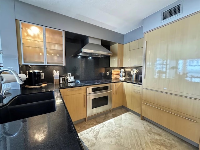 kitchen featuring wall chimney range hood, sink, backsplash, light brown cabinetry, and oven