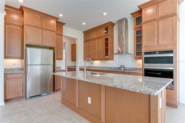 kitchen with wall chimney range hood, sink, appliances with stainless steel finishes, light stone counters, and an island with sink