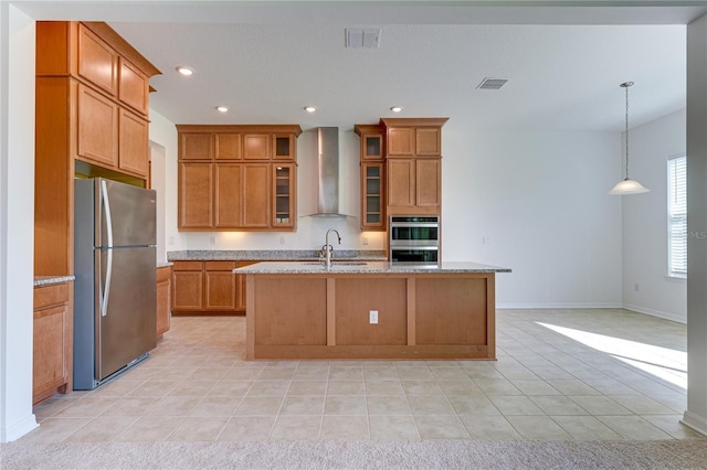 kitchen with pendant lighting, stainless steel appliances, light stone countertops, a center island with sink, and wall chimney exhaust hood