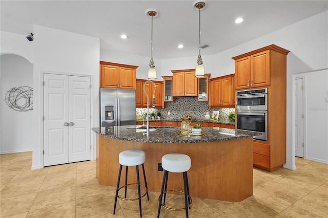 kitchen with appliances with stainless steel finishes, a center island with sink, brown cabinets, dark stone countertops, and glass insert cabinets