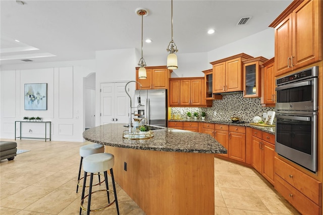 kitchen featuring visible vents, dark stone counters, glass insert cabinets, appliances with stainless steel finishes, and a kitchen island with sink