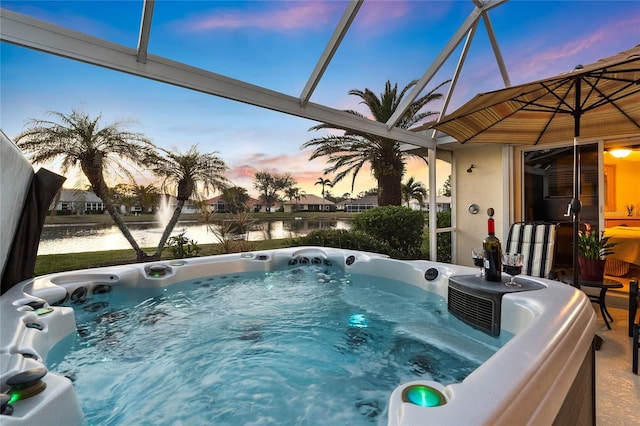 pool at dusk with a hot tub, a water view, and glass enclosure