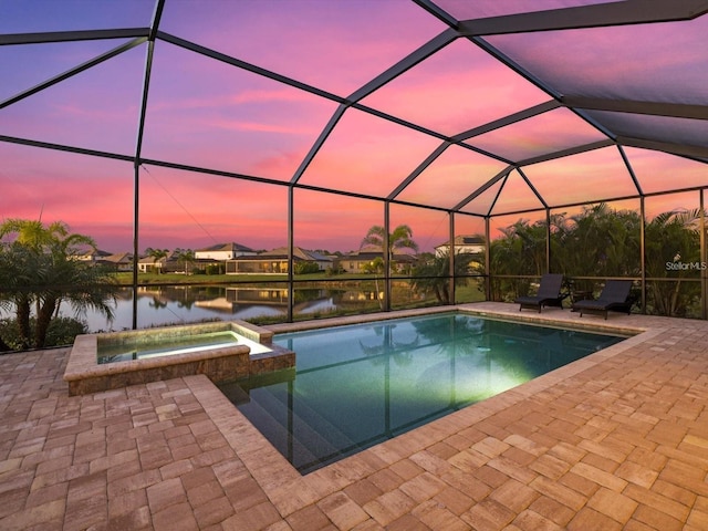 pool at dusk with an in ground hot tub, a water view, a patio area, and glass enclosure