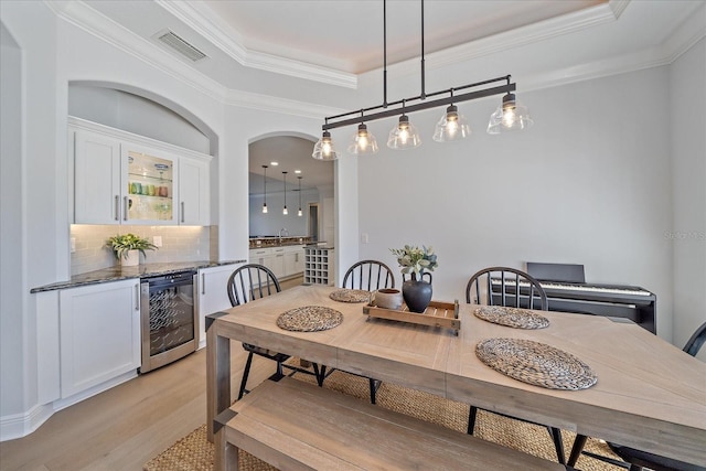 dining area featuring wine cooler, crown molding, bar area, light wood-type flooring, and a raised ceiling