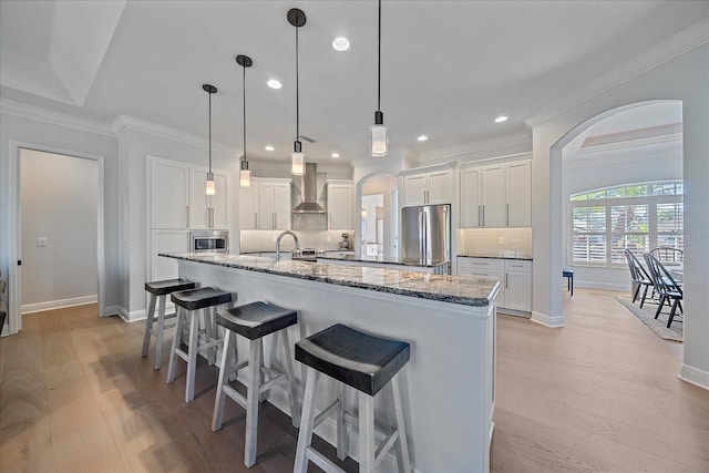 kitchen featuring a large island with sink, white cabinets, stainless steel appliances, and wall chimney exhaust hood