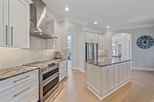 kitchen with white cabinetry, appliances with stainless steel finishes, wall chimney exhaust hood, and dark stone counters