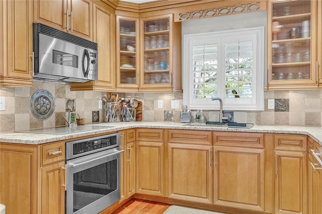 kitchen with stainless steel appliances, decorative backsplash, glass insert cabinets, a sink, and light stone countertops