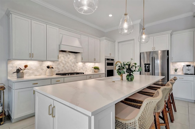 kitchen with premium range hood, appliances with stainless steel finishes, decorative light fixtures, white cabinets, and a kitchen island with sink