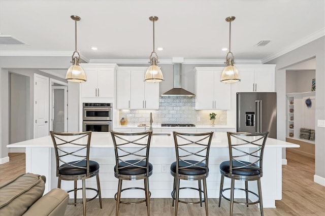 kitchen with wall chimney range hood, appliances with stainless steel finishes, white cabinetry, a center island with sink, and decorative light fixtures