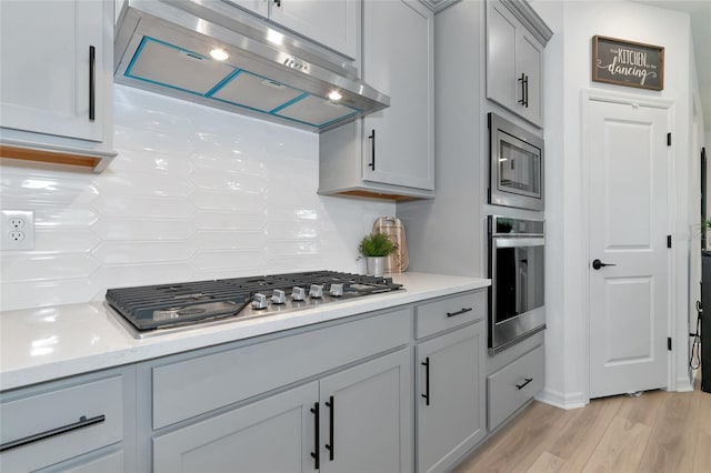 kitchen with light wood finished floors, tasteful backsplash, appliances with stainless steel finishes, gray cabinets, and under cabinet range hood