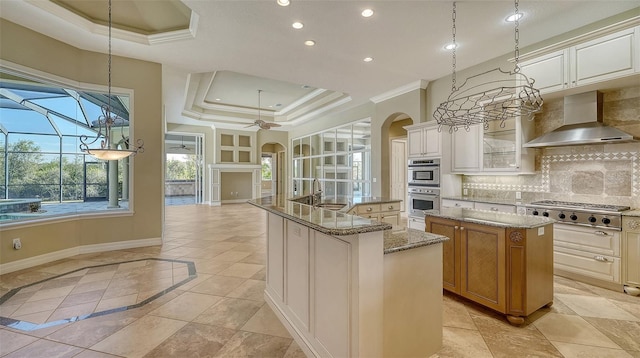 kitchen with an island with sink, light stone counters, stainless steel appliances, a raised ceiling, and wall chimney exhaust hood