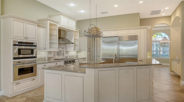 kitchen featuring wall chimney range hood, appliances with stainless steel finishes, a kitchen island with sink, backsplash, and decorative light fixtures