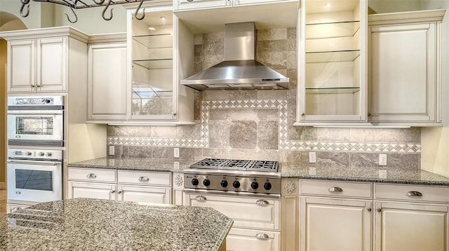 kitchen with backsplash, dark stone countertops, double oven, and wall chimney range hood