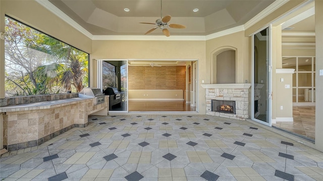 interior space featuring an outdoor stone fireplace, ornamental molding, light tile patterned floors, ceiling fan, and a tray ceiling