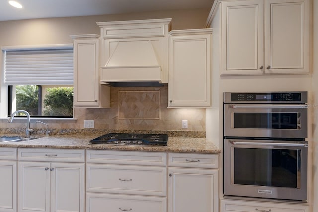 kitchen with sink, premium range hood, appliances with stainless steel finishes, light stone counters, and tasteful backsplash