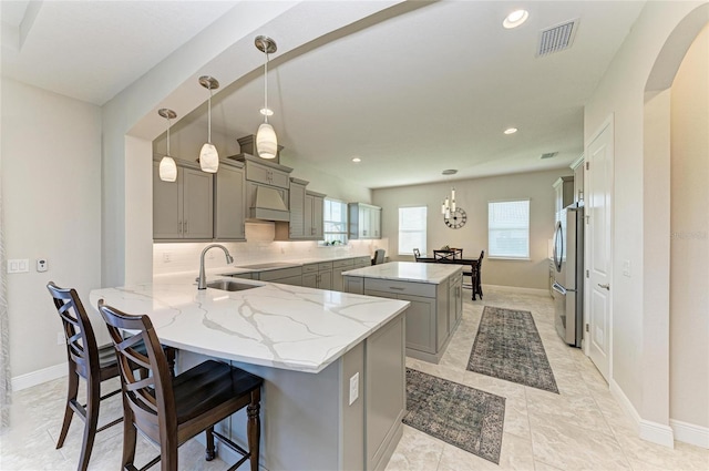 kitchen featuring hanging light fixtures, stainless steel fridge, gray cabinets, kitchen peninsula, and custom range hood