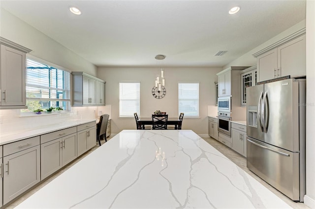 kitchen featuring pendant lighting, gray cabinets, appliances with stainless steel finishes, light stone countertops, and decorative backsplash