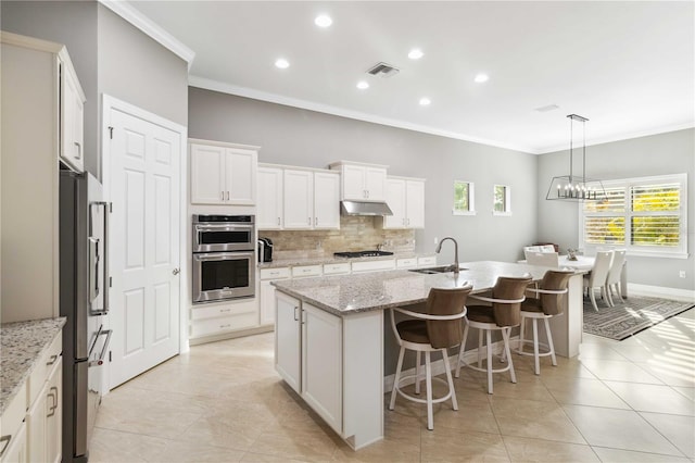 kitchen featuring appliances with stainless steel finishes, tasteful backsplash, an island with sink, white cabinets, and light stone countertops