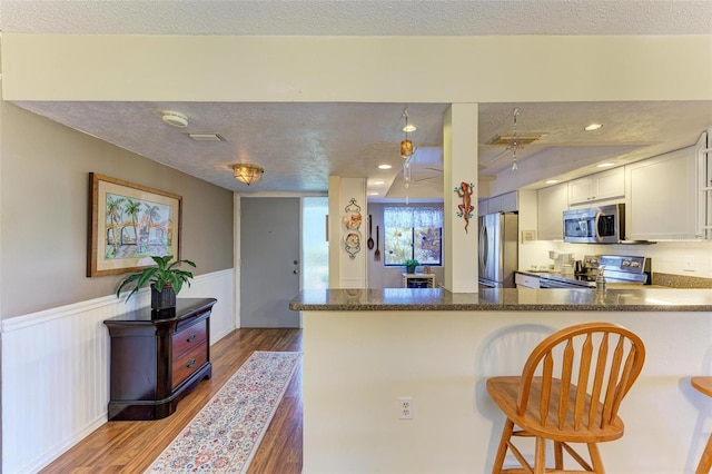 kitchen with a wainscoted wall, a peninsula, light wood-style flooring, stainless steel appliances, and a textured ceiling