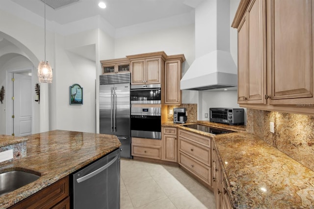 kitchen with wall chimney range hood, hanging light fixtures, stainless steel appliances, tasteful backsplash, and light stone countertops