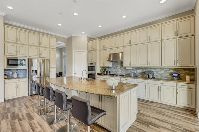 kitchen with cream cabinets, a breakfast bar area, stainless steel appliances, light stone countertops, and a center island with sink