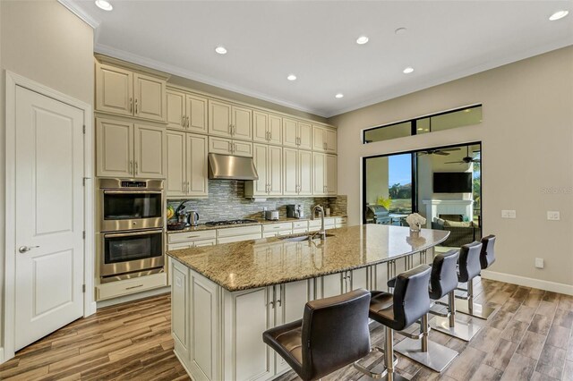 kitchen featuring appliances with stainless steel finishes, an island with sink, sink, light stone counters, and cream cabinets