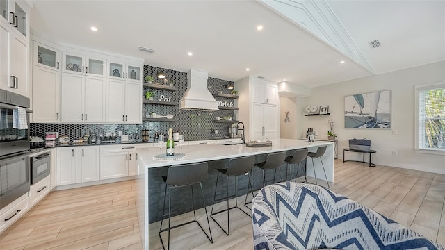 kitchen with lofted ceiling, a kitchen island with sink, custom range hood, and white cabinets