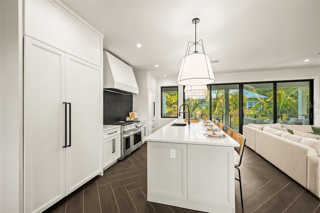 kitchen with sink, premium range hood, a kitchen island with sink, backsplash, and range with two ovens