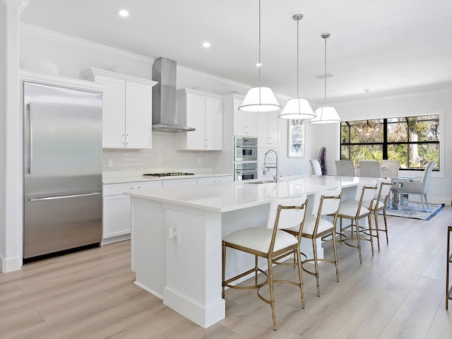 kitchen with white cabinetry, hanging light fixtures, a large island with sink, appliances with stainless steel finishes, and wall chimney range hood