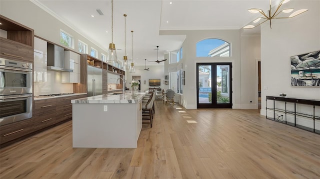 kitchen with wall chimney exhaust hood, decorative light fixtures, a center island with sink, light hardwood / wood-style flooring, and appliances with stainless steel finishes