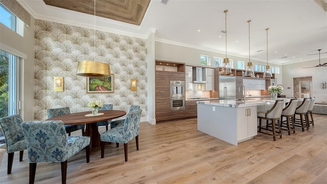 kitchen with appliances with stainless steel finishes, pendant lighting, an island with sink, light wood-type flooring, and wall chimney exhaust hood