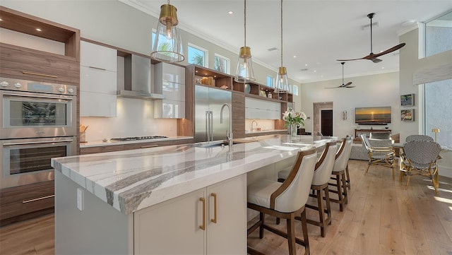 kitchen with wall chimney exhaust hood, decorative light fixtures, a center island with sink, stainless steel appliances, and white cabinets