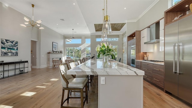 kitchen with wall chimney exhaust hood, crown molding, a center island with sink, appliances with stainless steel finishes, and light hardwood / wood-style floors