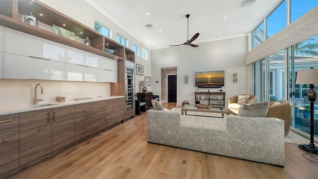 living room featuring sink, light hardwood / wood-style flooring, ceiling fan, a high ceiling, and ornamental molding