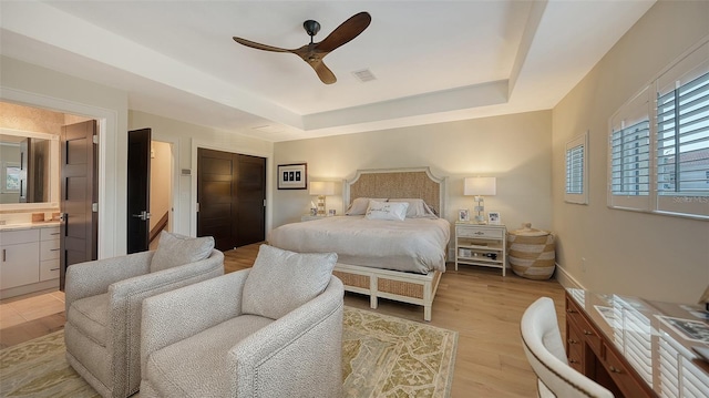 bedroom with ceiling fan, connected bathroom, a tray ceiling, a closet, and light wood-type flooring