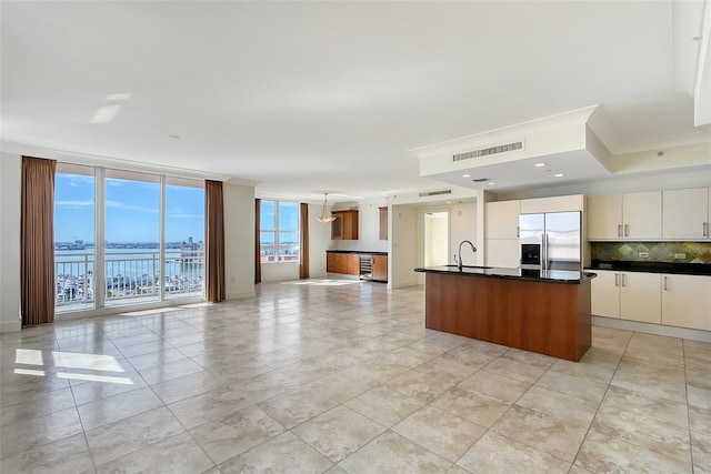 kitchen with stainless steel refrigerator with ice dispenser, a water view, white cabinetry, a kitchen island with sink, and decorative backsplash