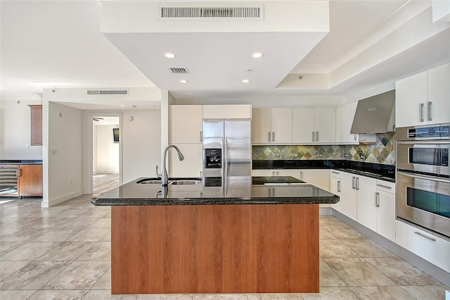 kitchen featuring appliances with stainless steel finishes, an island with sink, sink, white cabinets, and wall chimney exhaust hood